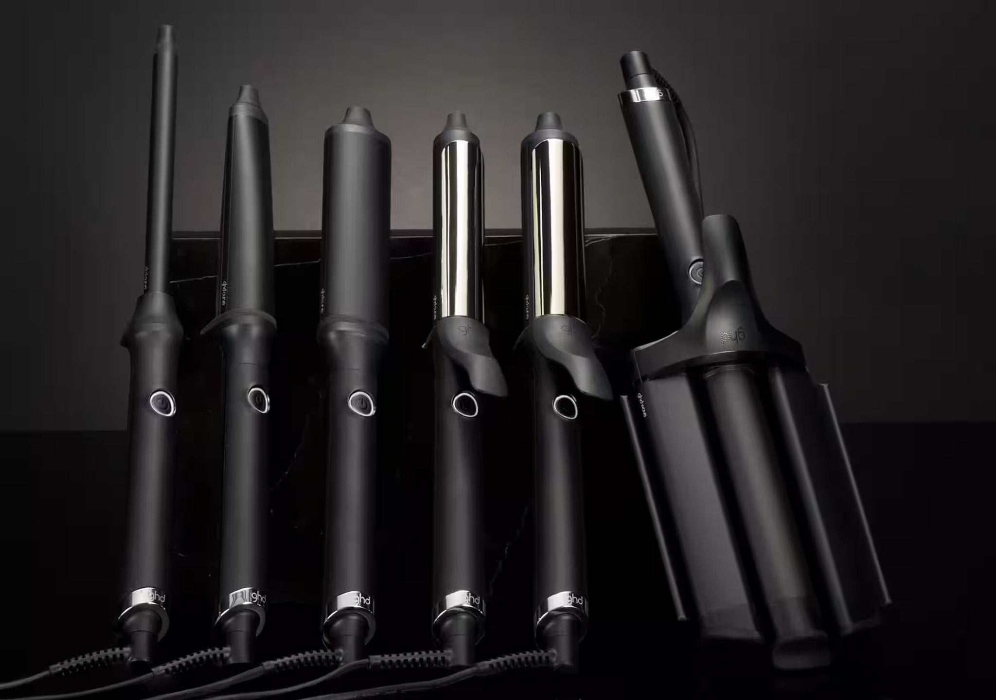 A row of black curling irons, wands, flatirons leaning against a black background.