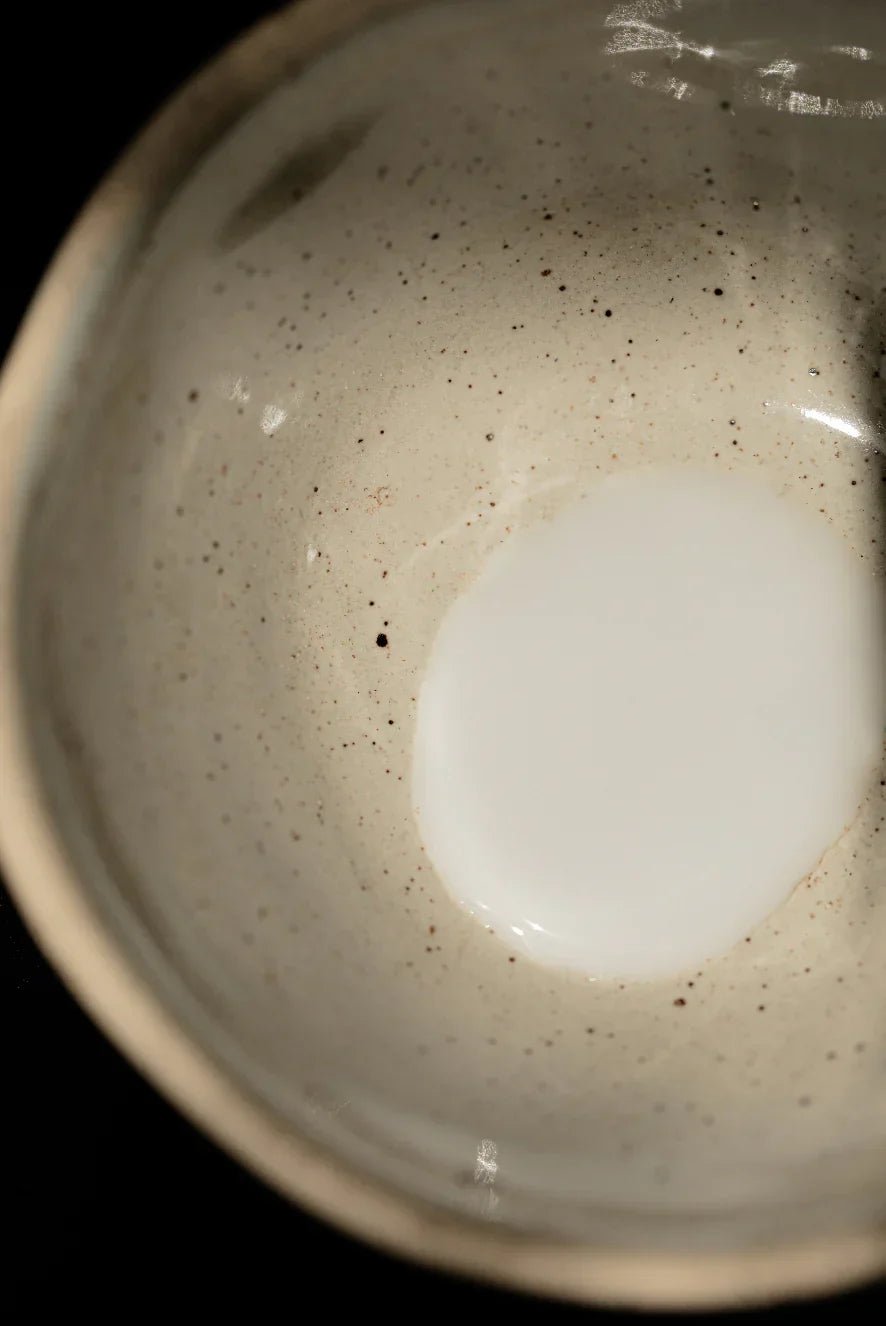 A small amount of white liquid sits at the bottom of a beige, speckled ceramic bowl against a dark background.