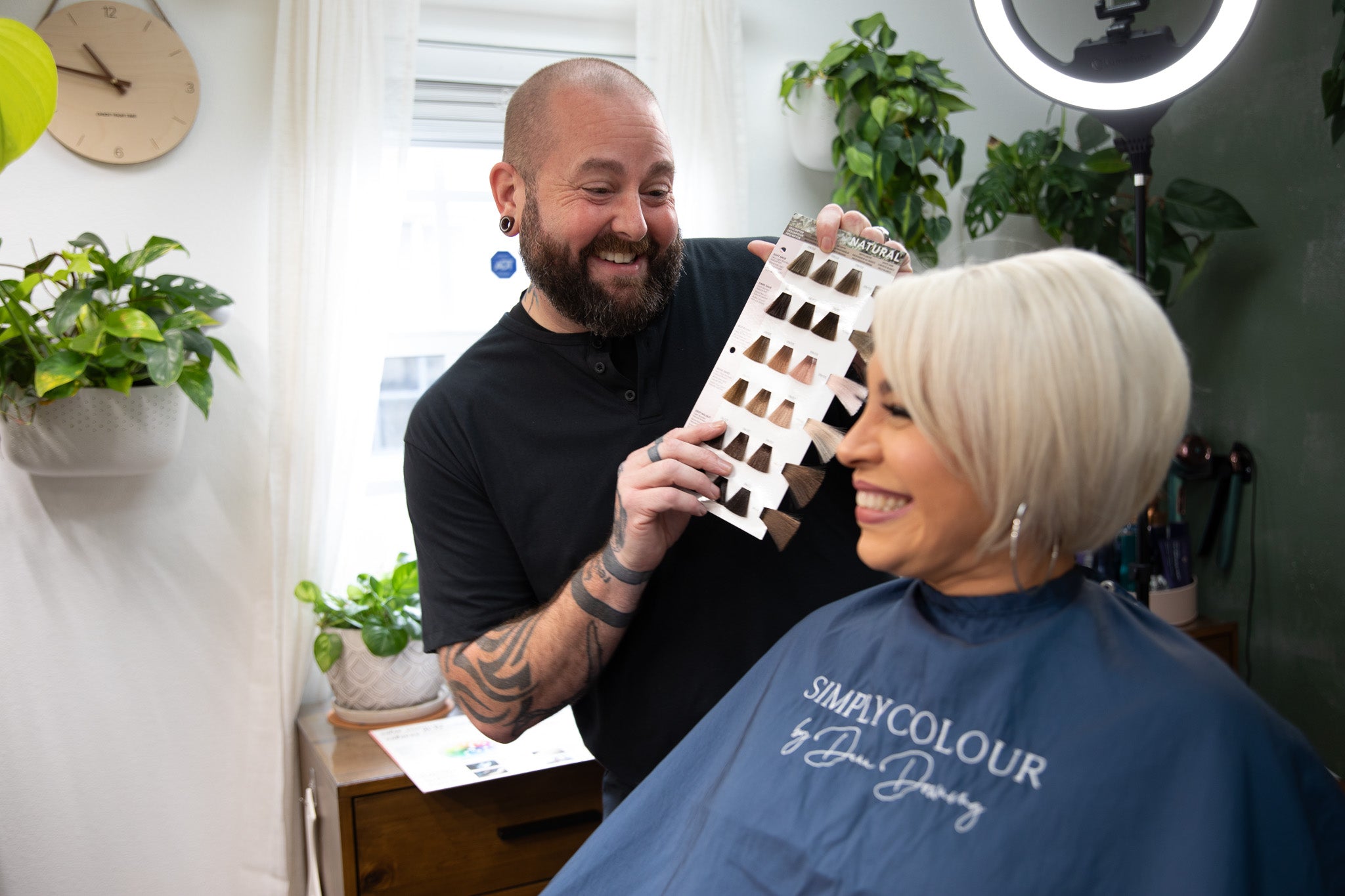 A hairstylist holds up a hair color sample chart and smiles at a client with short, platinum blonde hair, who is seated and wearing a salon cape. They are in a well-lit salon with plants in the background.