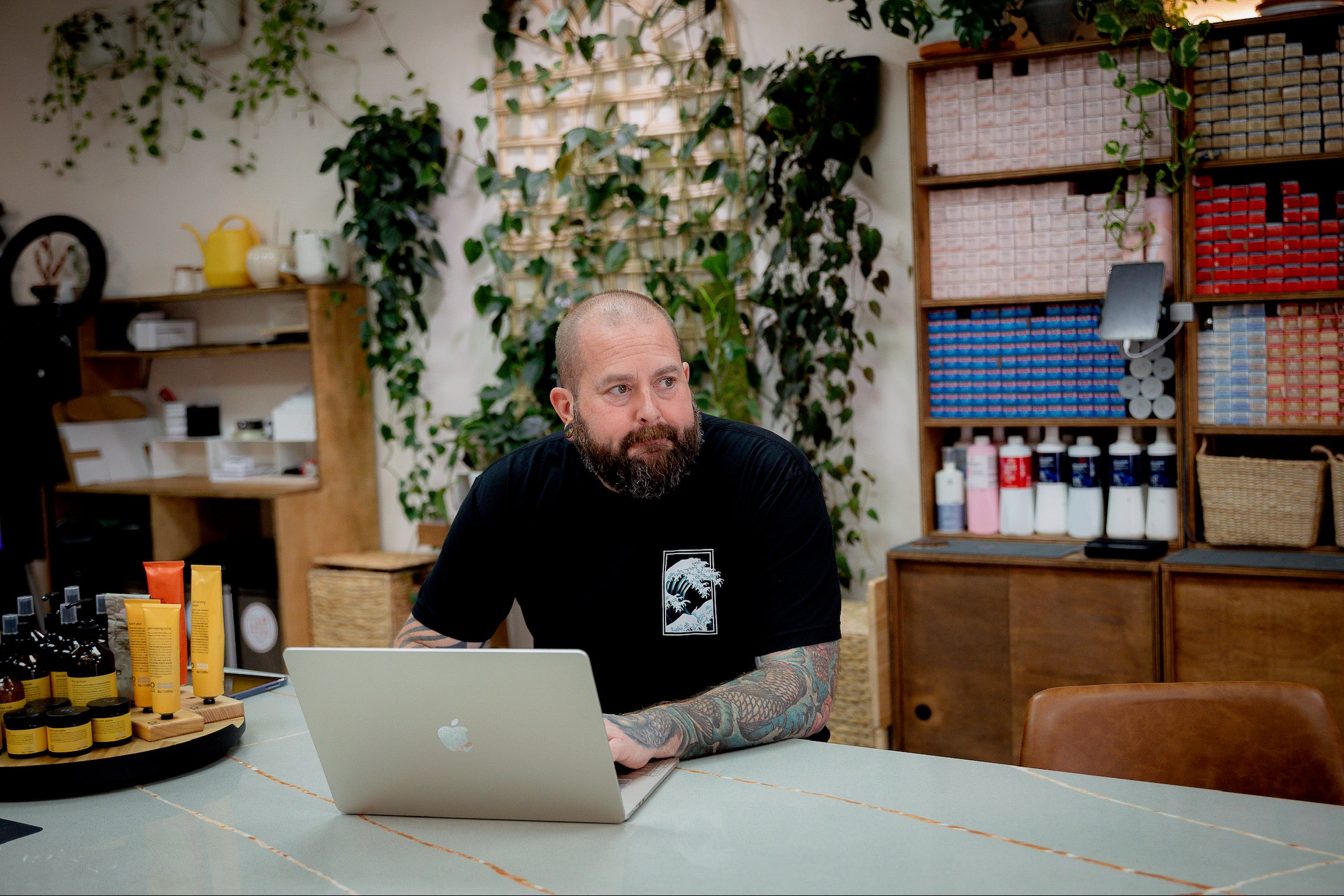 Dean Downing, owner of Simply Colour, sitting at the salon counter with a laptop. A color bar and wooden furniture and plants are in the background.