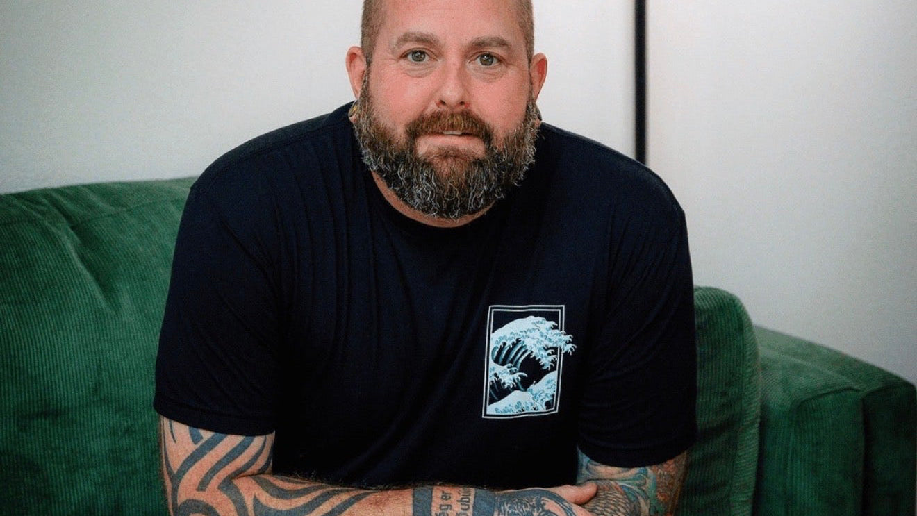 Dean Downing, owner of SImply Colour, a man with tattoos, in a black shirt and green pants sitting on a green couch