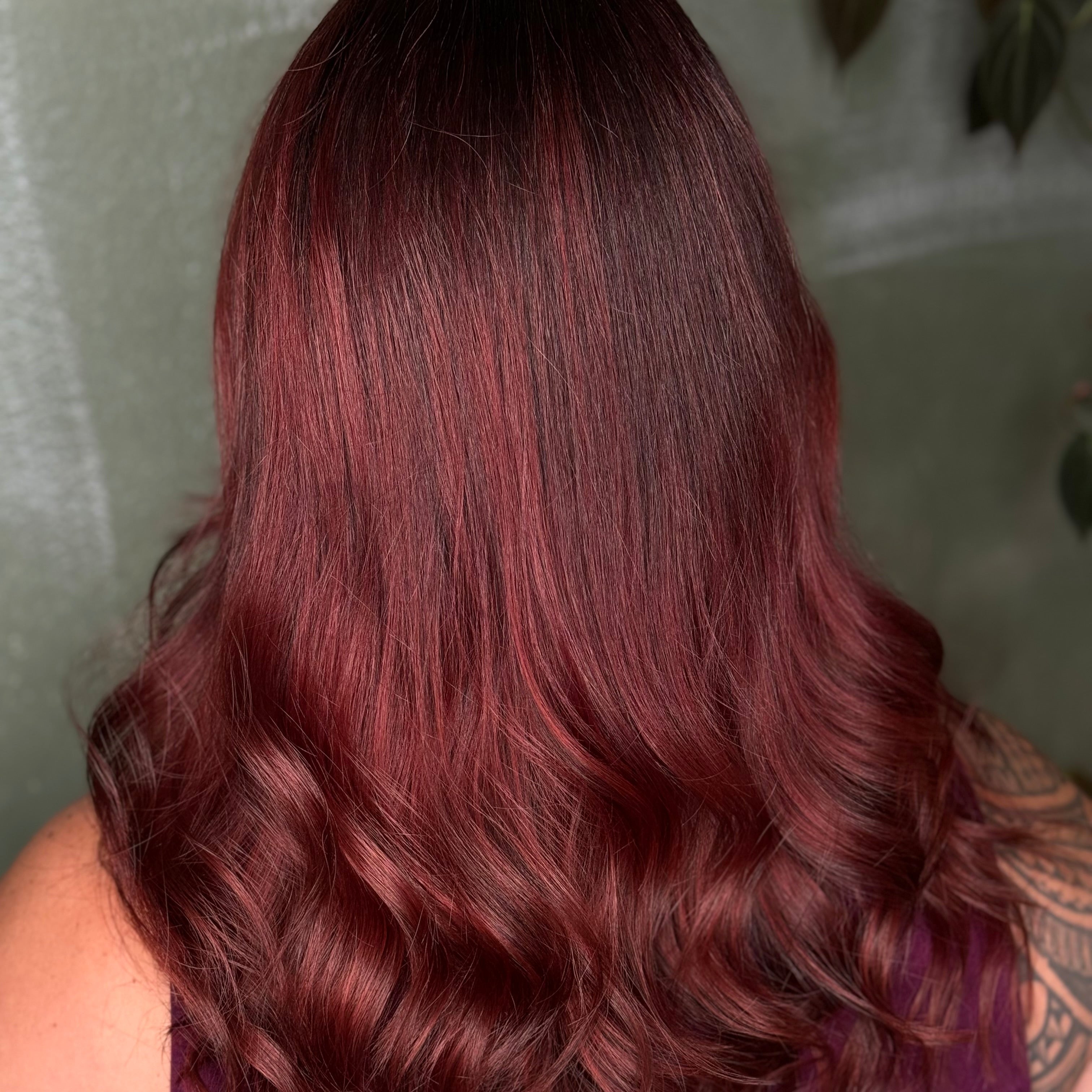 A person with long, wavy hair dyed a deep burgundy red is seen from behind. The hair appears shiny and healthy, and part of a tattooed shoulder is visible. The background is blurred green with some leaves.