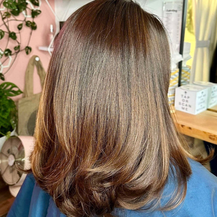 A person with medium-length, light brown hair sits in a salon chair wearing a blue cape. Shelves with hair products and potted plants are visible in the background, along with a mirror on the wall.