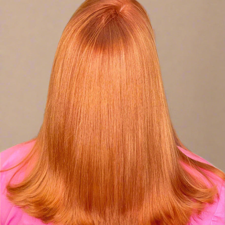 Person with straight, shoulder-length, vibrant copper hair facing away from the camera, wearing a pink top in a salon setting.