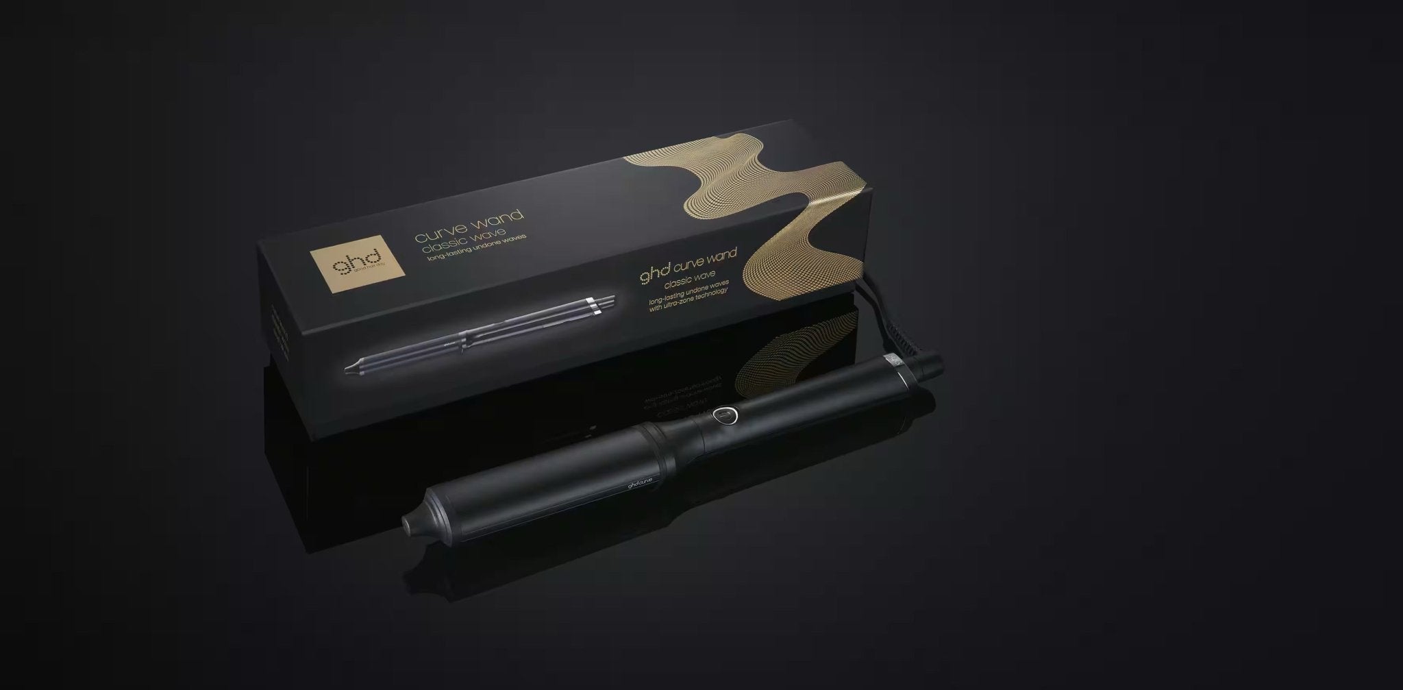 The GHD CLASSIC WAVE - OVAL CURLING WAND by GHD, featuring a ceramic-coated barrel and power cord, sits before its elegant box with gold wave designs—ideal for creating glamorous Hollywood waves against a dark background.