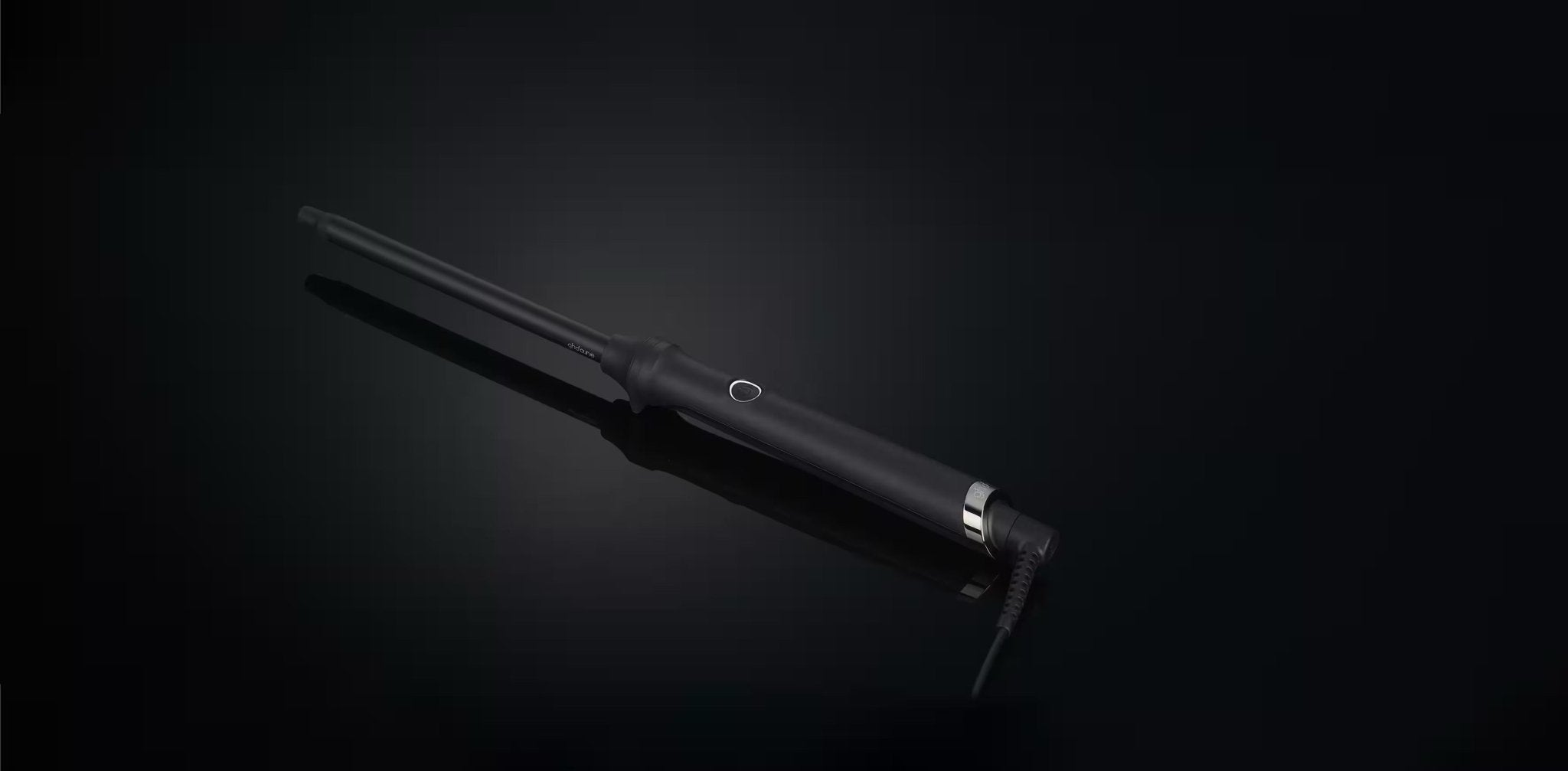 The GHD THIN WAND - 0.5" Curling Wand in black by GHD, featuring a slim barrel and single button, rests diagonally on a dark surface with its power cord at the bottom right—ideal for creating defined curls.