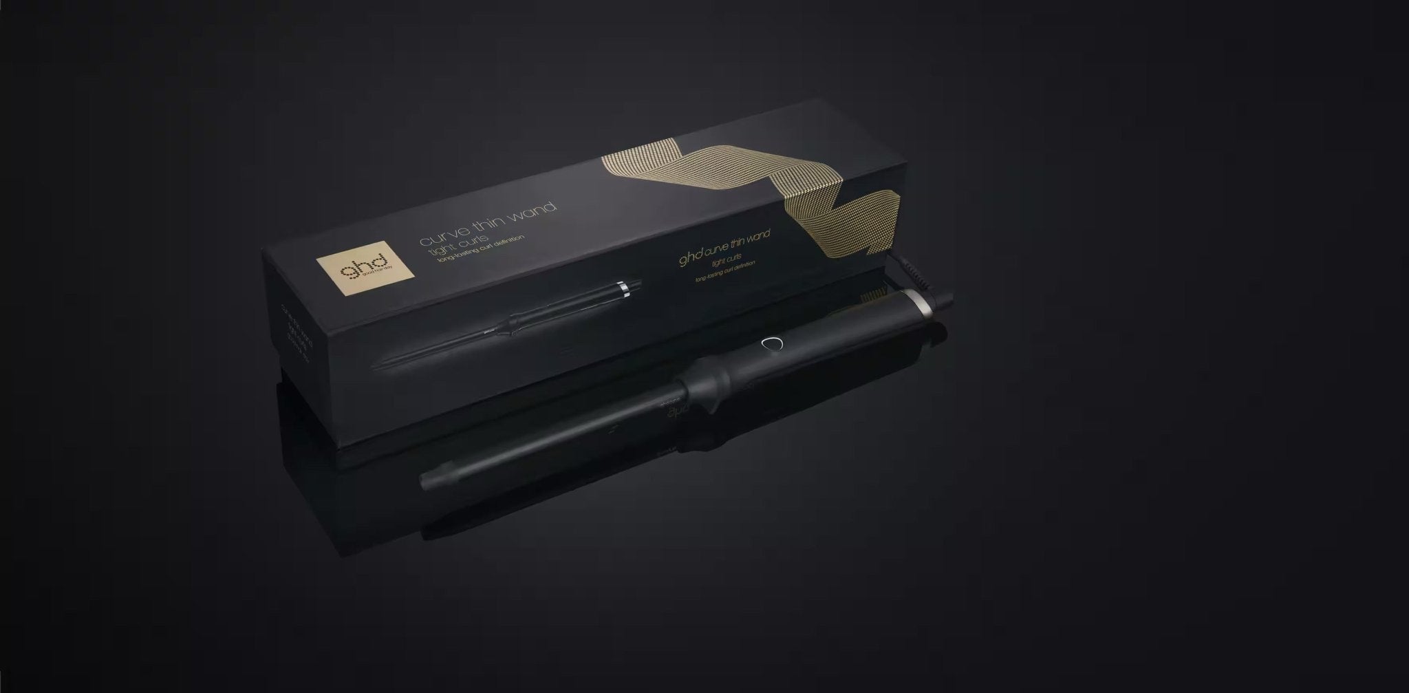 A GHD THIN WAND 0.5" CURLING WAND in black sits before its sleek black and gold box, both labeled by GHD, on a reflective surface—ideal for creating defined curls with this ultra-thin curling wand.