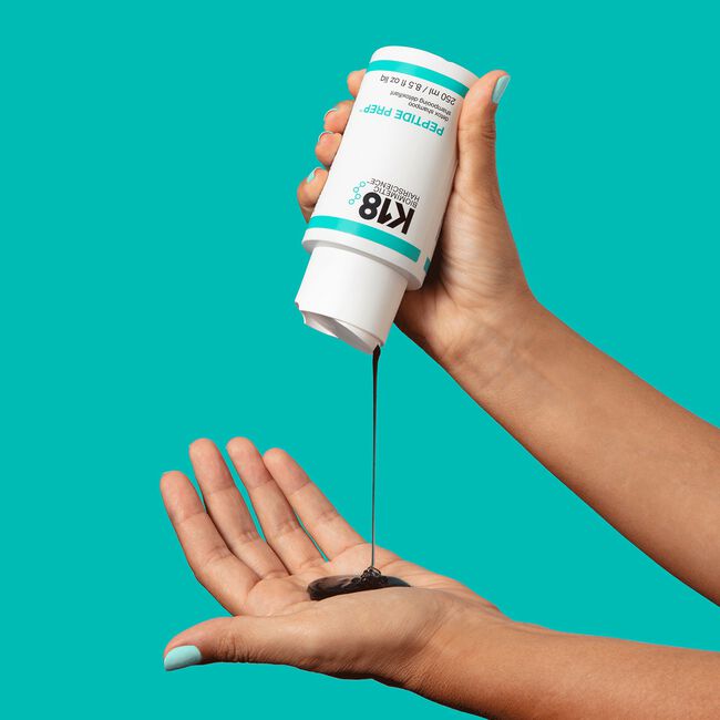Hand holding a K18 product with another hand catching the liquid on a teal background