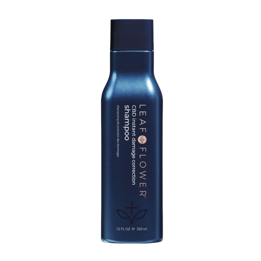 A dark blue bottle of Leaf and Flower Damage Correction Shampoo with white and pink text stands upright on a beige background. This color-safe, frizz-controlling formula is available in multiple sizes from Leaf and Flower.