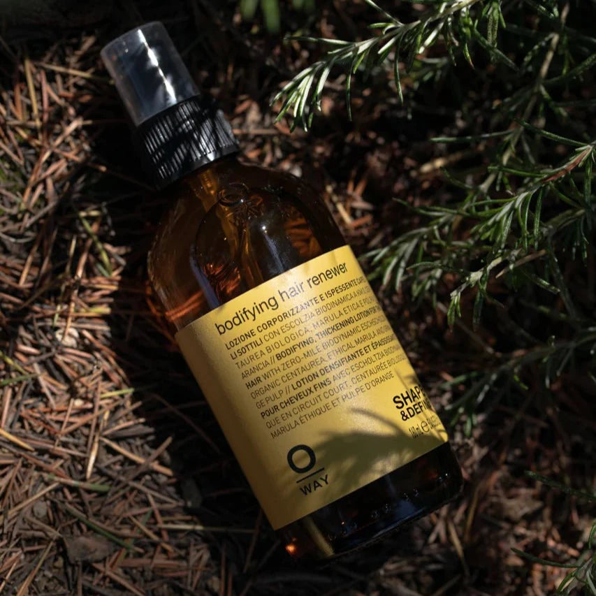 A brown Oway Bodifying Hair Renewer spray bottle rests among pine needles and greenery, partly shaded—an anti-hair fall densifying spray for fine hair that boosts volume naturally.