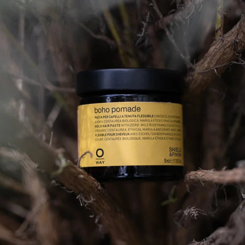 A black Oway Boho Pomade jar with a yellow label rests among brown twigs. This texturizing cream pomade offers a flexible hold and laid-back matte finish, featuring biodynamic marrubium. The label includes text in several languages.