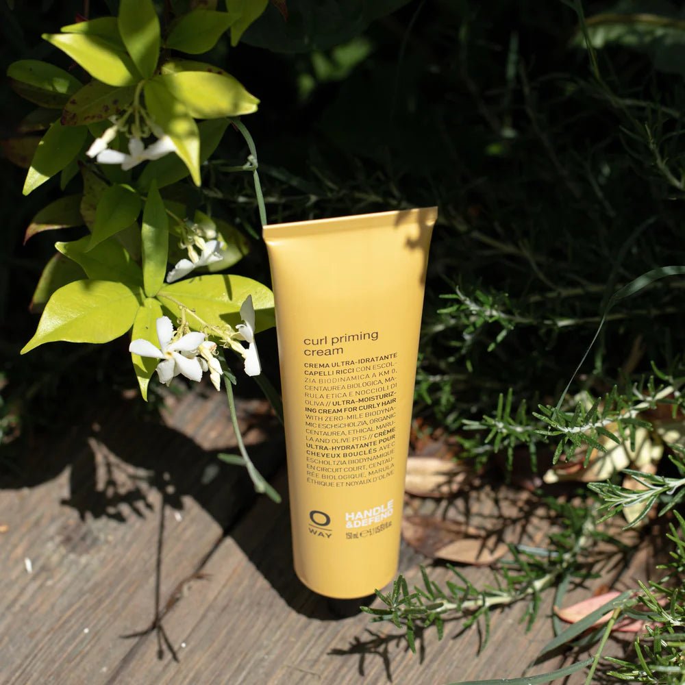 A yellow tube of Oway Curl Priming Cream | Moisture-Rich Curl Primer for Frizz-Free Styling stands upright on a wooden surface amid green foliage and small white flowers, bathed in sunlight. Crafted with Marula Oil, it nourishes and defines curls.