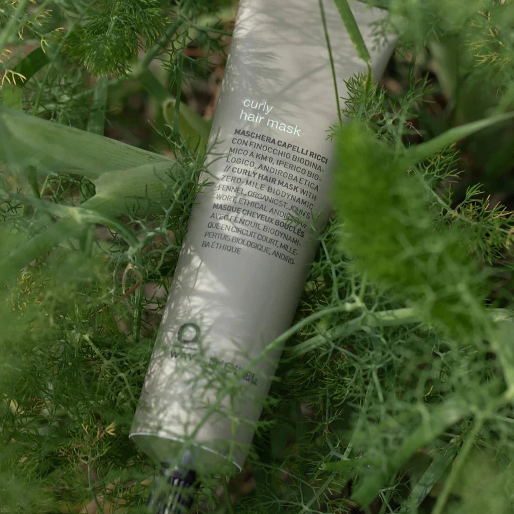 A green Oway Curly Hair Mask tube, labeled "Definition and Moisture Mask for Natural Curls," rests among leafy plants. Its black print is partially hidden by foliage, reflecting its deep moisture and curl-defining benefits.