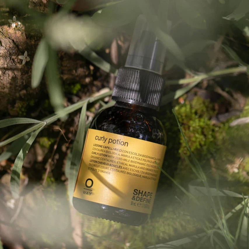 An Oway Curly Potion Curl-Defining Serum with a yellow label and spray nozzle rests on green moss and grass outdoors, partly surrounded by leaves and illuminated by natural light.