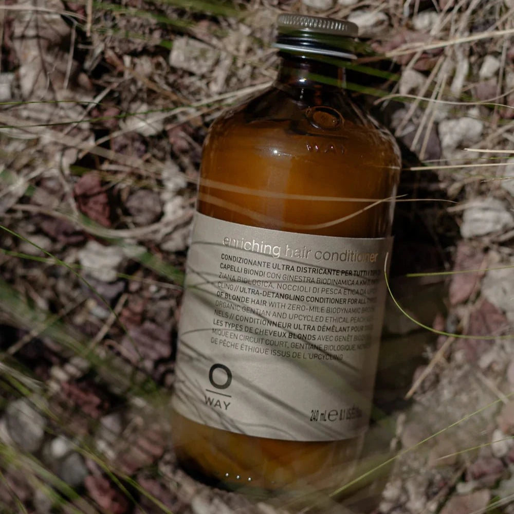 A brown glass bottle labeled "Oway Enriching Hair Conditioner | Ultra Detangling For Dry Brittle Strands" lies on rocky ground with dry grass, offering intense detangling and restored shine for dry, brittle hair.