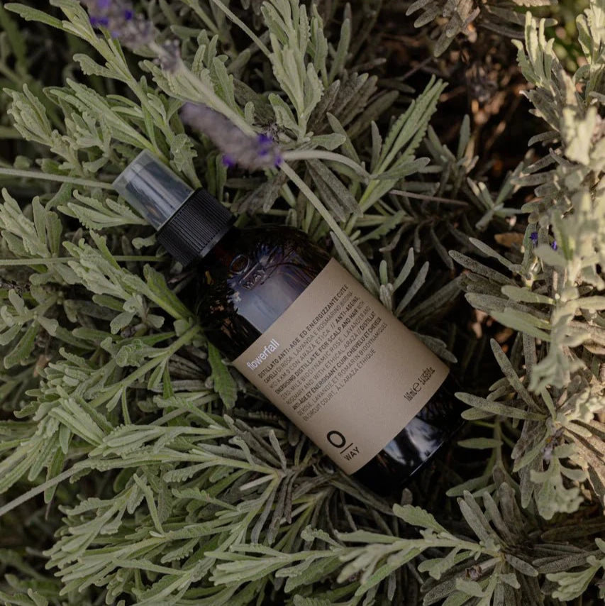 A brown bottle labeled "Oway Flowerfall | Aromatherapeutic Scalp and Mind Balancing Mist" sits on lush green lavender foliage. The product is from Oway, designed to balance scalp and mind with its aromatherapeutic formula.