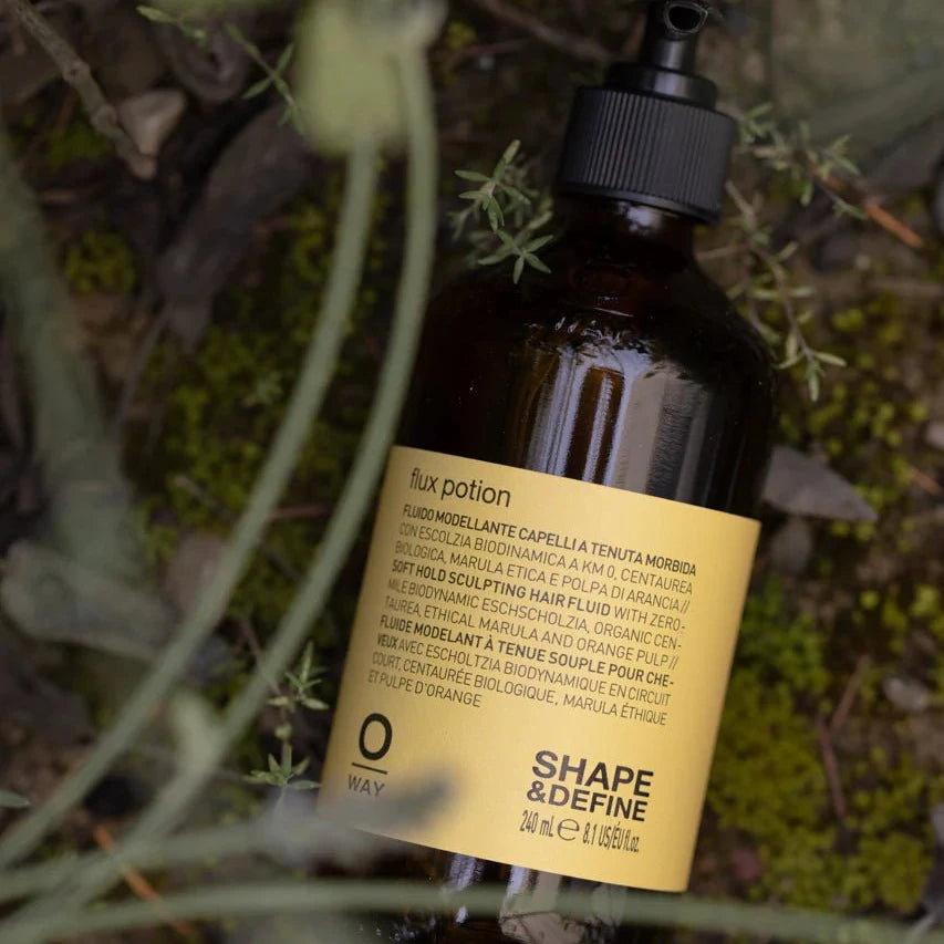 A brown Oway Flux Potion Lightweight Volumizing Serum bottle with a gold label and black pump rests on moss, surrounded by green plants. The label features the Oway logo and mentions Biodynamic Marrubium for soft body and hold.