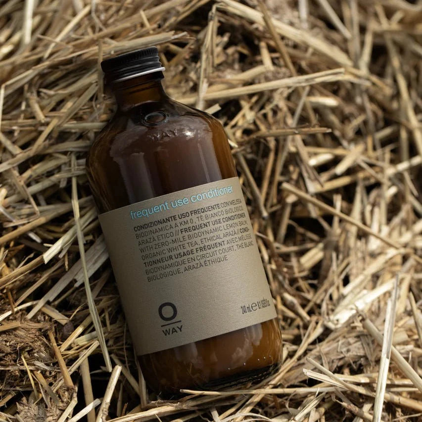 A brown glass bottle of Oway Frequent Use Conditioner | Lightweight Conditioner for Everyday Use rests on straw. Its label, in multiple languages, states it's for all hair types and contains 240 ml.