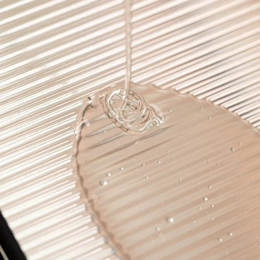 A close-up of Oway Frequent Use Hair & Scalp Bath, a gentle daily shampoo for all hair types, being poured onto a ridged, light-colored surface, forming a small puddle with visible droplets around it.