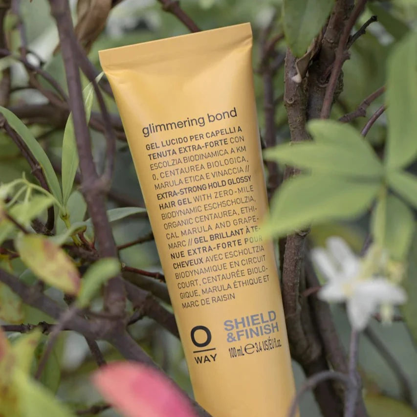 A yellow Oway Glimmering Bond tube—Bonding Shine Treatment for Damaged, Dull Hair—stands upright among green leaves and white flowers. The label displays product details in both Italian and English.