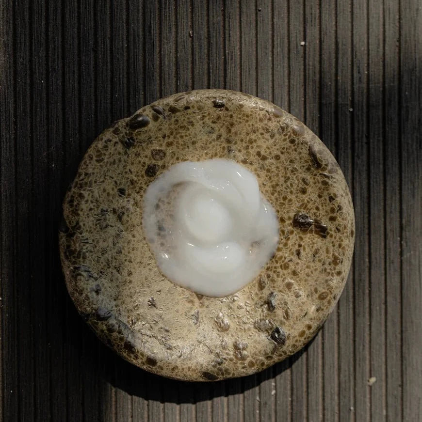 A touch of Oway Next Day Cream | Soft Styling Cream for Next-Day Texture Revival is elegantly swirled atop a textured, round brown stone on a striped surface, capturing the essence of Oway’s next-day texture revival.