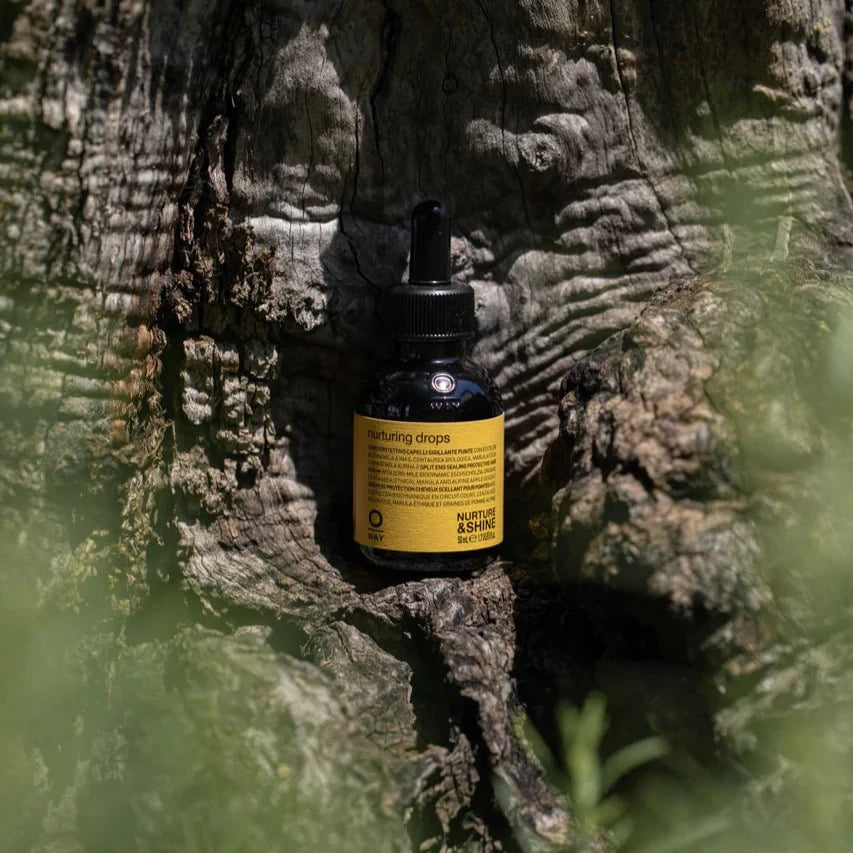 A small bottle of Oway Nurturing Drops | Silkening Serum for Split Ends and Heat Protection stands upright against textured tree bark, its yellow label visible amid green foliage in the foreground.