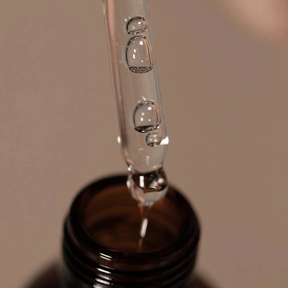 A close-up of a glass dropper with clear liquid and bubbles held above the Oway Nurturing Drops | Silkening Serum for Split Ends and Heat Protection bottle by Oway, set against a neutral background.