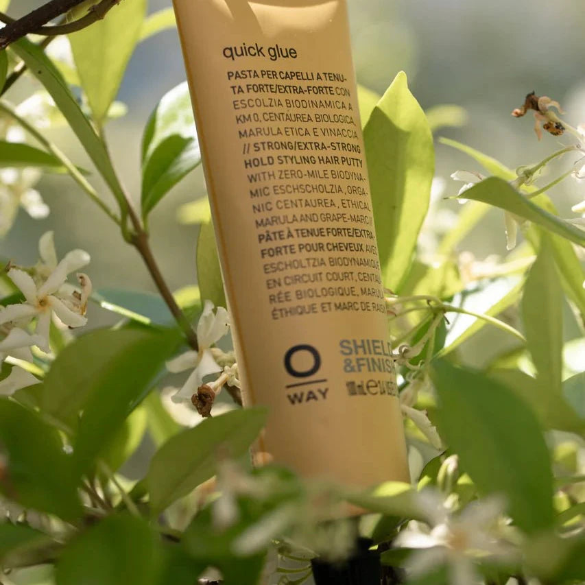 A gold tube of Oway Quick Glue | Ultra-Hold Styling Glue for Creative Hair Sculpting stands upright among green leaves and white flowers, with sculpting instructions in several languages and product details near the bottom.