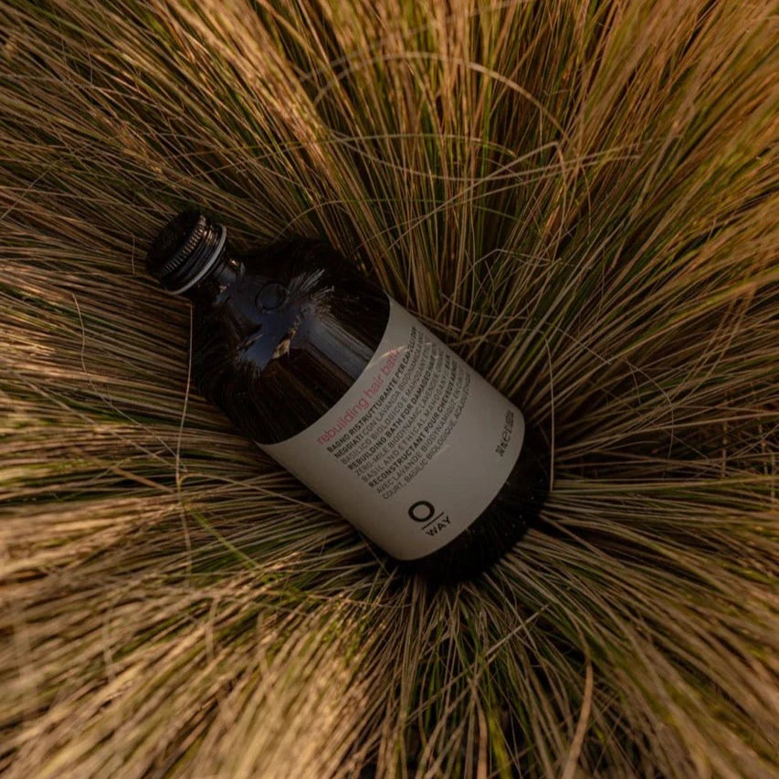 A brown Oway Rebuilding Hair Bath bottle with a white label lies on its side in dry grass. The label shows it's a Strengthening Shampoo for brittle, weak hair, suggesting repair and revitalization benefits.