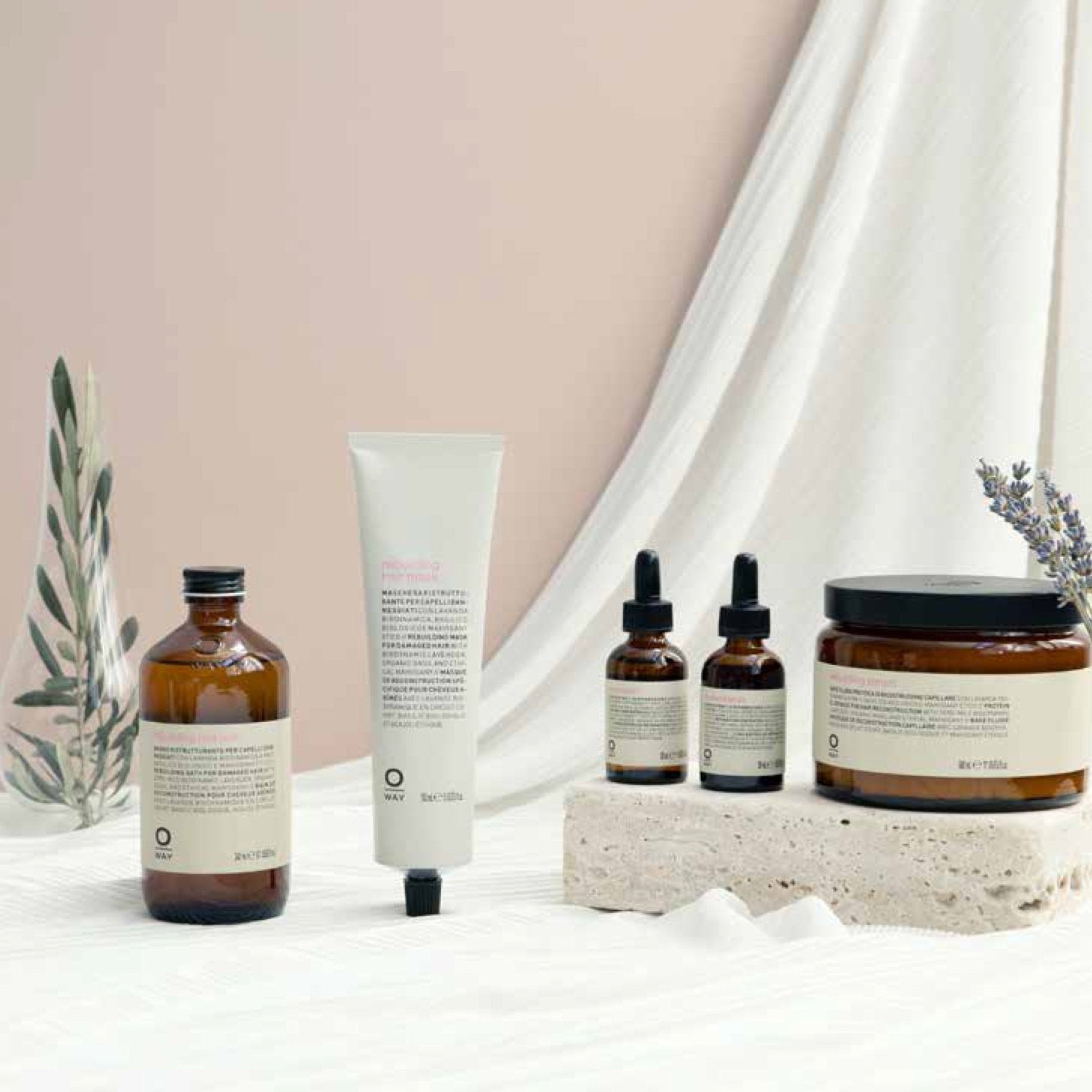 An assortment of Oway skincare in amber and white bottles, jars, a tube, and droppers—plus Oway Rebuilding Hair Bath Strengthening Shampoo for brittle hair—rests on light fabric with sprigs of greenery and lavender.
