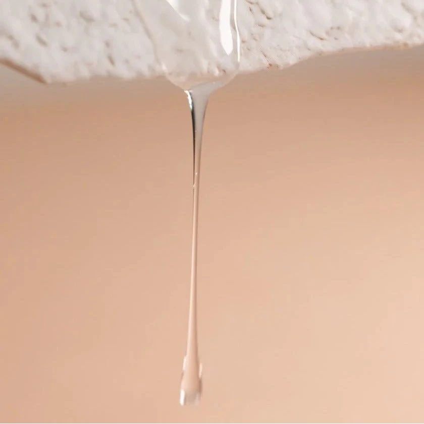 A close-up of a clear liquid drip hanging from a textured white surface above a beige background, evoking the purity and renewal found in Oway Rebuilding Hair Bath, the strengthening shampoo for brittle, weak hair.
