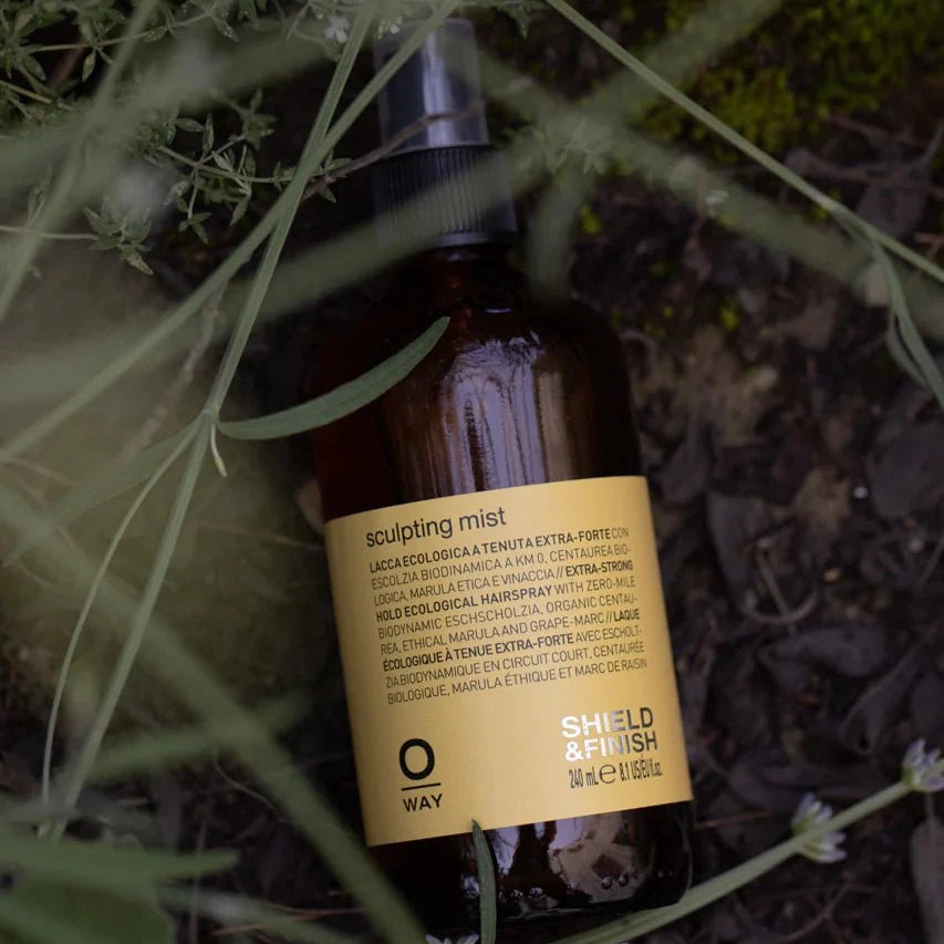 A brown glass Oway Sculpting Mist bottle, labeled "Long-Lasting Finishing Spray with Flexible Hold," rests among green leaves and grass. The label highlights its "SHIELD & FINISH" flexible hold formula.
