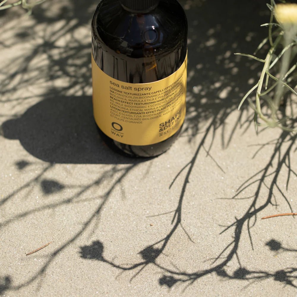 A brown bottle of Oway Sea Salt Spray | Beachy Texture Spray for Tousled Waves rests on sandy ground with plant shadows, featuring a yellow label with text and a black cap.