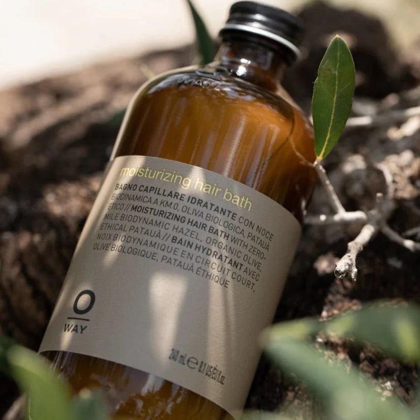 A brown Oway Moisturizing Hair Bath bottle labeled "Hydrating Shampoo for Dry, Lackluster Hair" sits on a natural surface outdoors, partly framed by green leaves. The multilingual label highlights its moisturizing benefits.