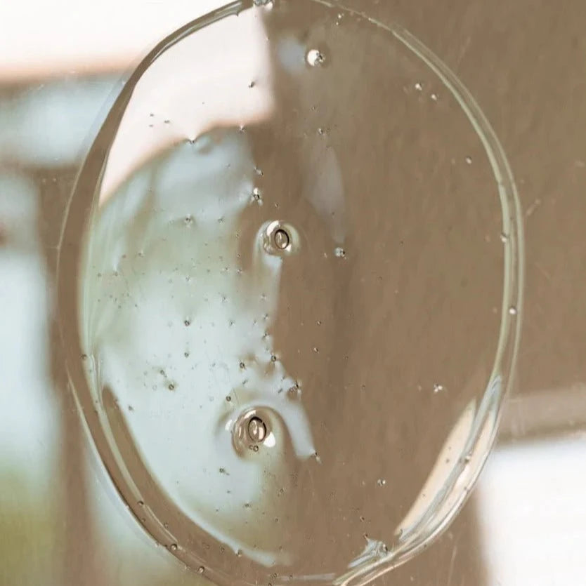 A close-up of Oway Moisturizing Hair Bath, a clear hydrating shampoo with air bubbles, spread in a circle on a smooth surface. The soft, blurred background highlights its nourishing formula for dry, lackluster hair by Oway.