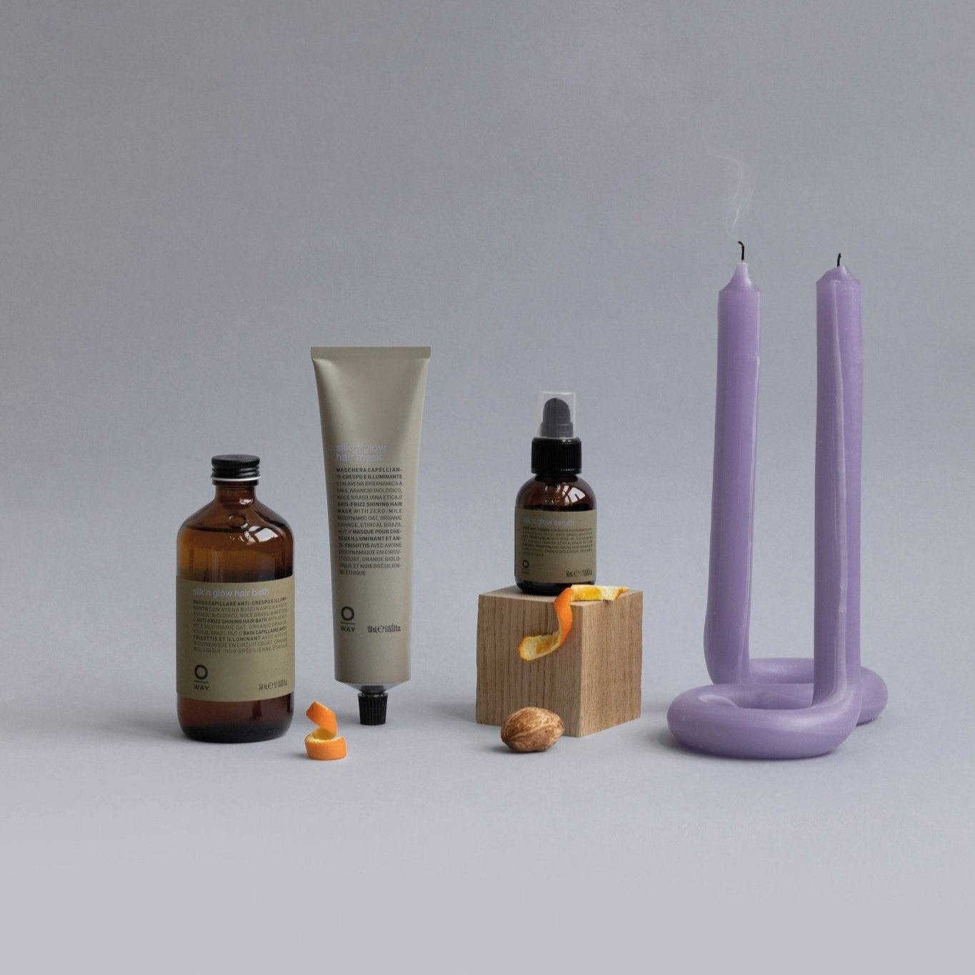 Three brown Oway Silk’N Glow Hair Bath containers, a wooden block, and two purple candles rest beside a nut and orange peel strips—an elegant scene evoking the nourishing power of Oway’s taming hair bath for frizz-prone hair.