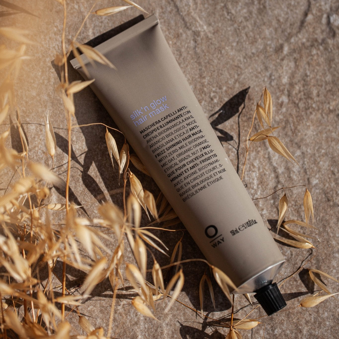 A tan tube labeled "Oway Silk’N Glow Hair Mask" with black text and a black screw cap rests on textured stone amid dried beige plants. Infused with Organic Cupuaçu Butter, this nourishing mask is ideal for dry, frizzy hair.