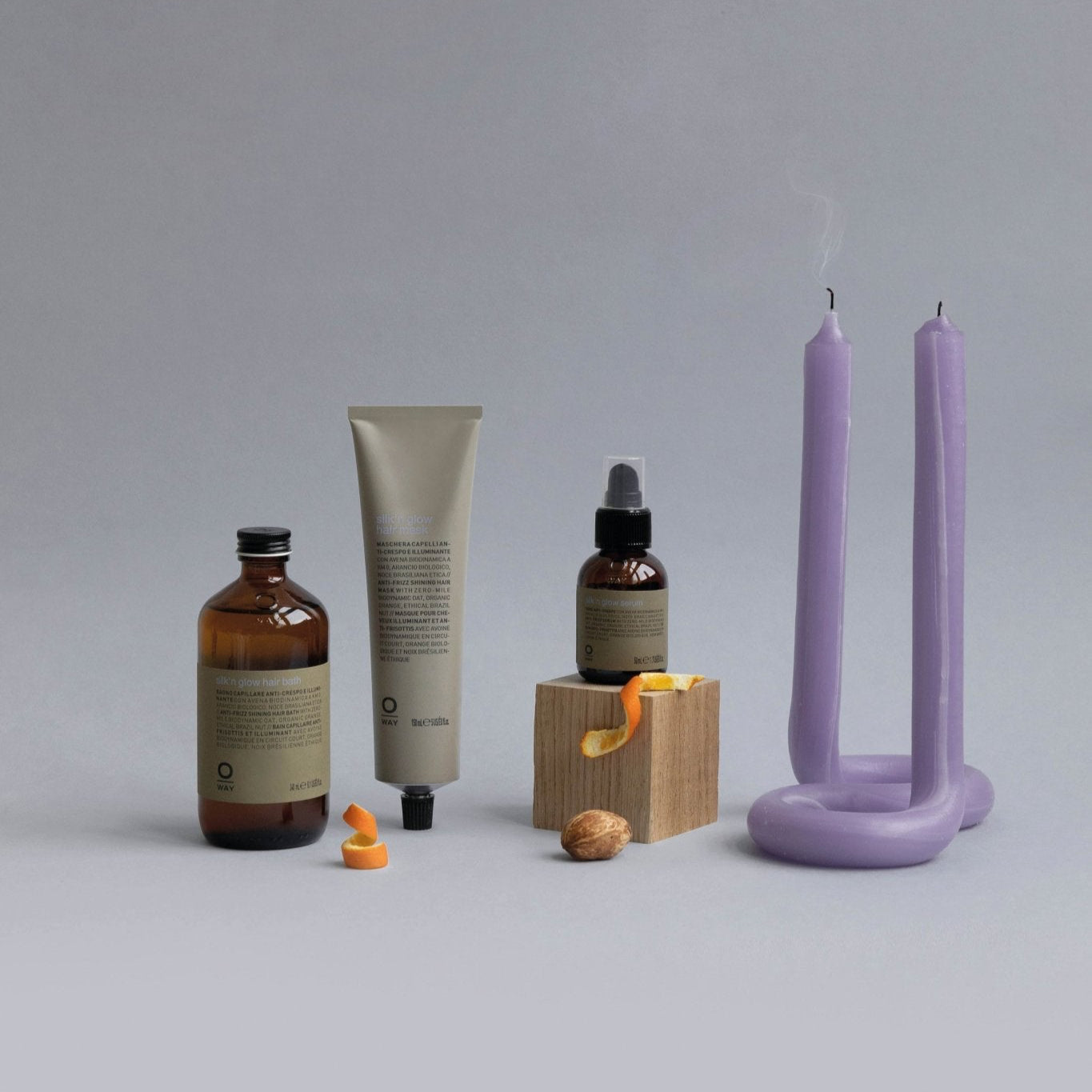 Three Oway Silk’N Glow Hair Mask products for dry, frizzy hair are arranged with a wooden block, orange peel, walnut, and purple candle on a grey background. Smoke wafts from the candle’s extinguished wicks.