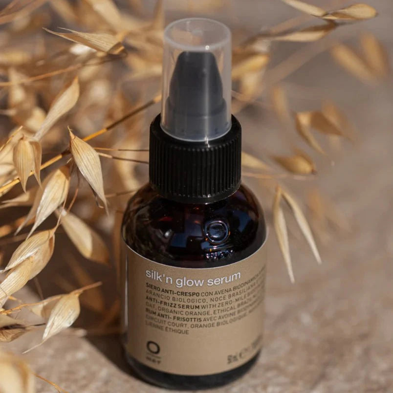 A brown glass bottle of Oway Silk’N Glow Serum | Anti-Frizz Shine Serum for Dry, Frizzy Hair with a pump dispenser rests on a beige surface beside dried wheat stalks; its label features nourishing Brazil nut oil and product details.