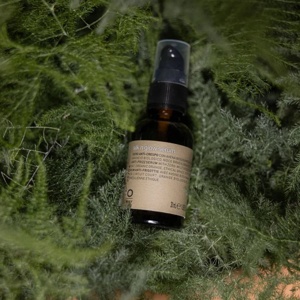 A brown glass dropper bottle labeled “Oway Silk’N Glow Serum | Anti-Frizz Shine Serum for Dry, Frizzy Hair” rests on dense green foliage. The Oway product features a black cap and is designed to add shine and tame frizz.