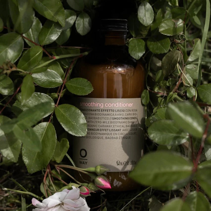 A brown Oway Smoothing Conditioner bottle with a black cap and white label sits among green leaves and a pink flower. Enriched with baobab oil, this detangling conditioner helps tame frizz for soft, polished hair.