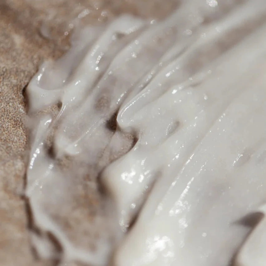 A close-up of Oway Smoothing Cream | Blow Dry Cream for Sleek, Straight Styles by Oway, creamy white and spread on a rough brown surface—ideal for taming frizzy hair and achieving smooth, straight looks.