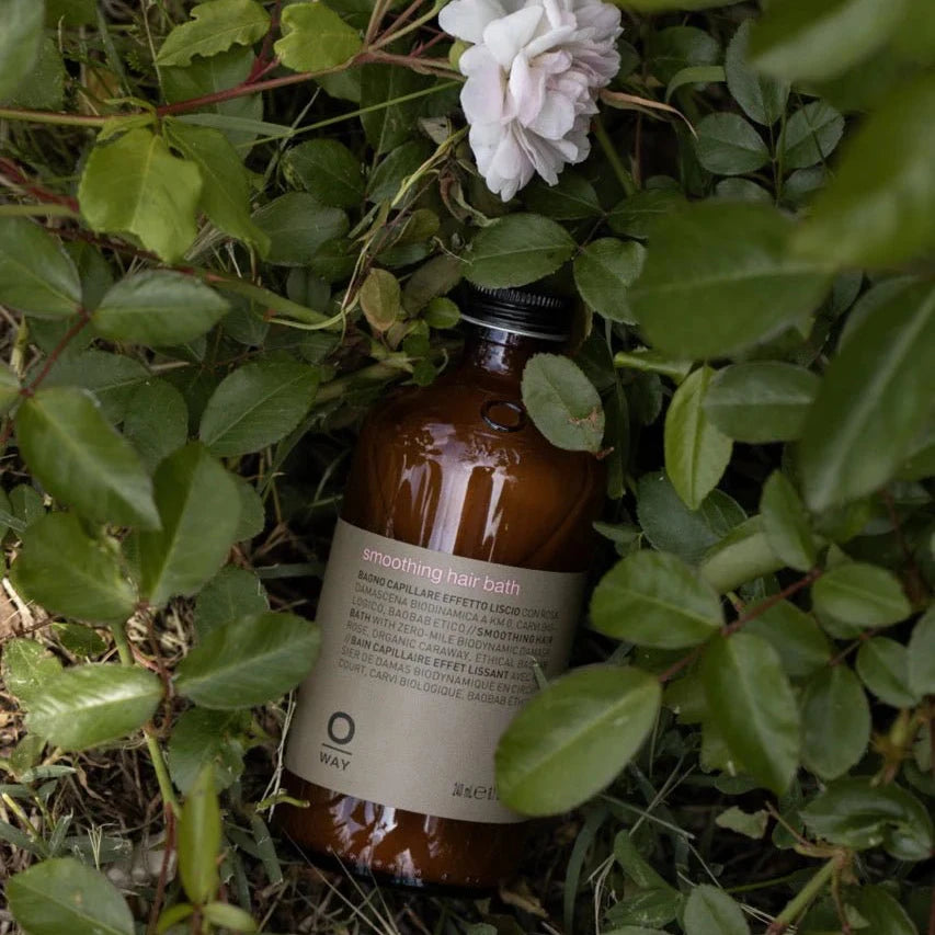 A brown Oway Smoothing Hair Bath bottle with a black cap and beige label rests among green leaves and a white rose with a pink bud, offering frizz control and nourishment for coarse or unruly hair.
