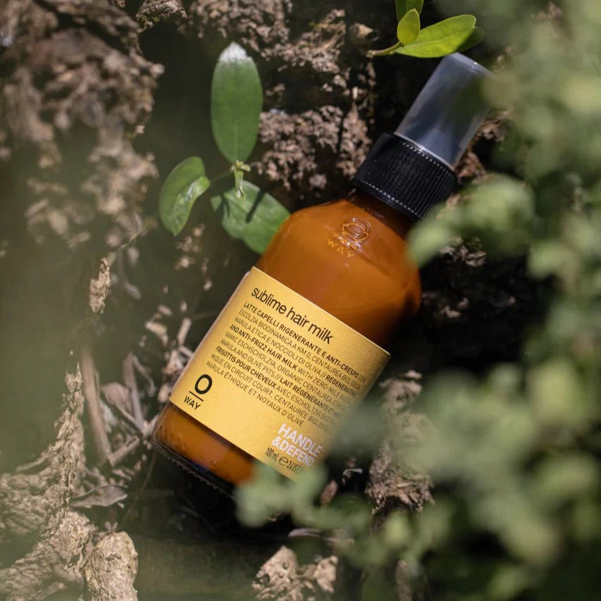 A brown bottle of Oway Sublime Hair Milk | Lightweight Leave-In Moisture Milk for Softness and Shine with a pump top stands among green plants and tree bark, photographed outdoors in natural light.