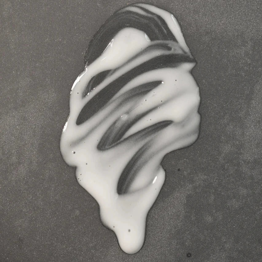 A small puddle of white liquid with streaks sits on a smooth, gray surface—resembling Oway Sublime Hair Milk, a lightweight leave-in moisture milk by Oway that delivers softness and shine while controlling frizz.