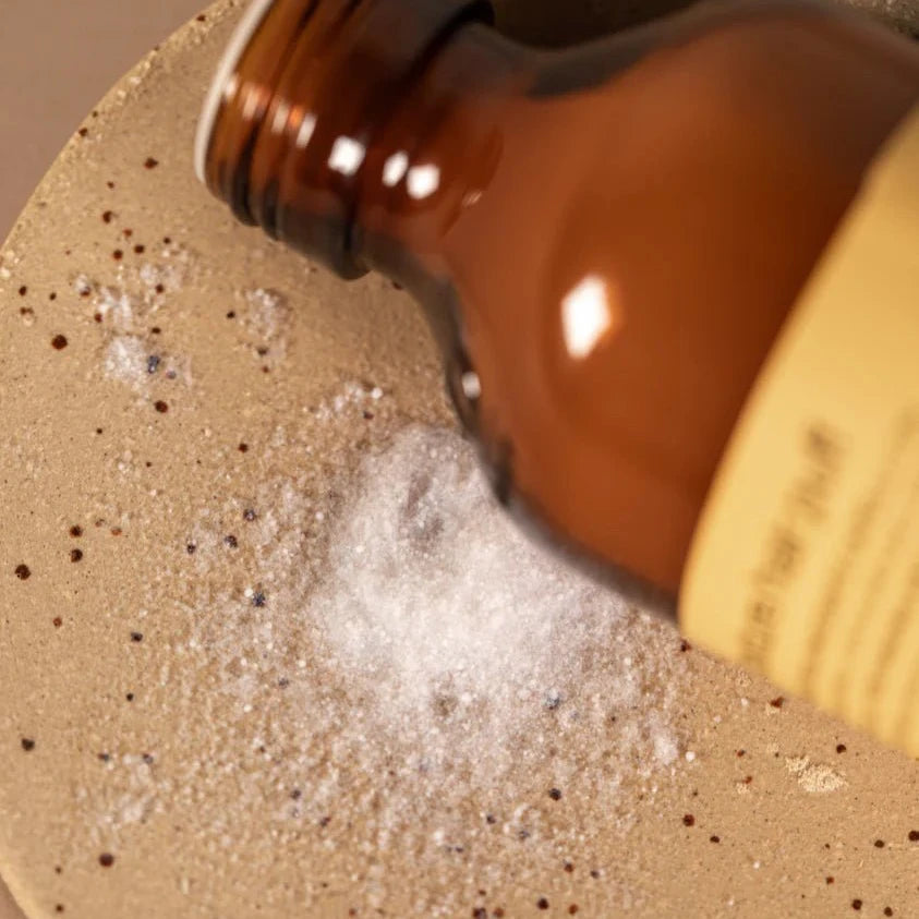 A brown glass bottle with a yellow label is tipped over a beige speckled surface, releasing Oway Volume and Texture Hair Puff—Volumizing Styling Powder for Instant Lift and texture by Oway.