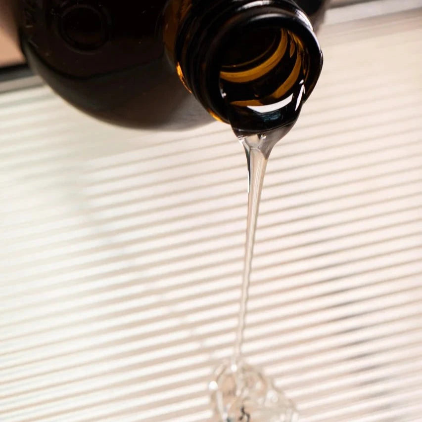 A close-up of Oway Volumizing Hair Bath, a gentle volumizing shampoo for fine hair, being poured from a dark brown glass bottle onto a white ridged surface. The organic ginger-infused liquid forms a small pool below.