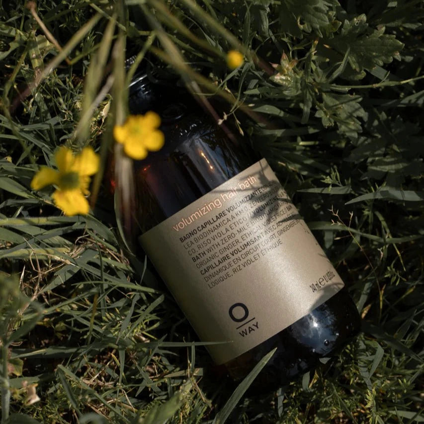 A brown bottle of Oway Volumizing Hair Bath, a gentle volumizing shampoo for fine or limp hair, rests on green grass with leaves and yellow flowers, sunlight casting shadows across the beige label.
