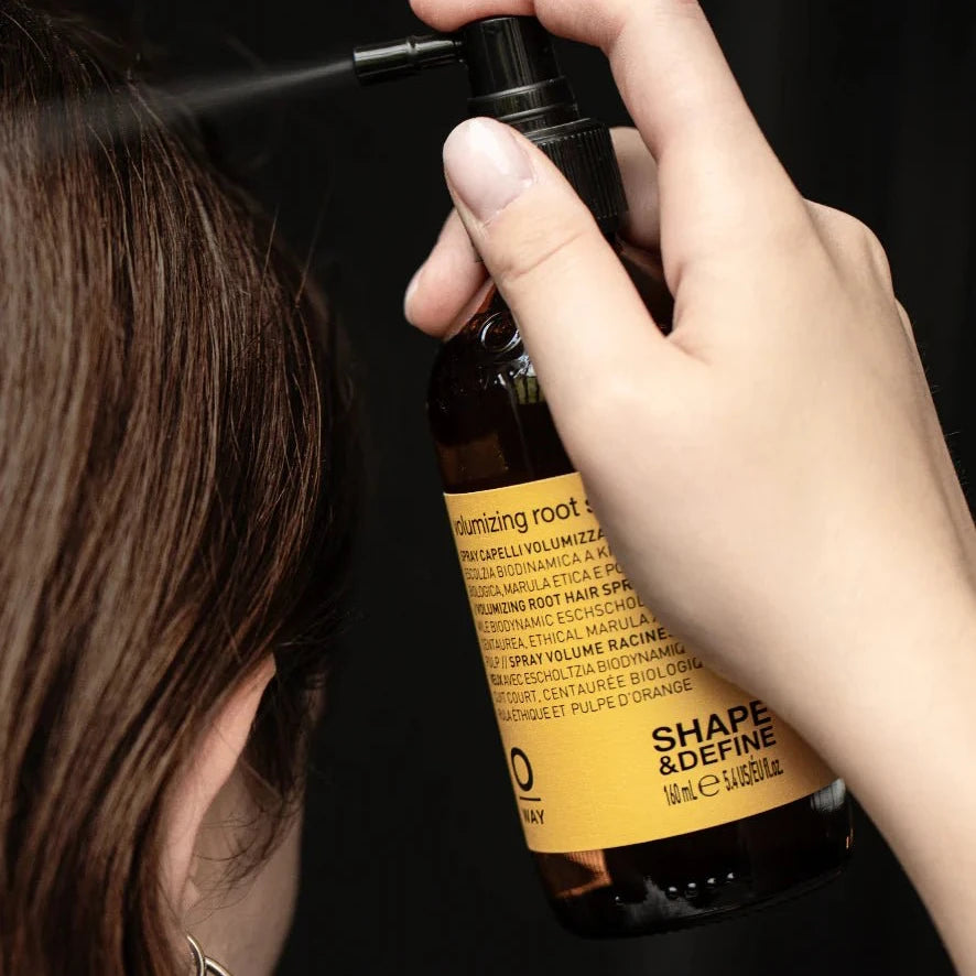 A person sprays Oway Volumizing Root Spray | Lightweight Root Lift for Fine, Flat Hair onto their hair, holding the brown bottle with their head turned away from the camera against a plain black background.