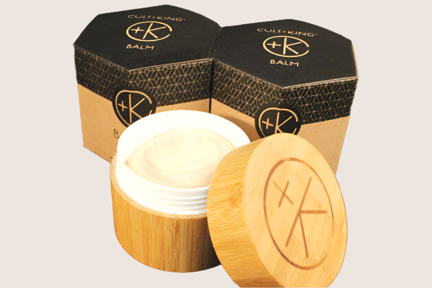 A round jar with a wooden lid holds Cult and King BALM | Hair to Toe, a creamy, moisturizing balm. It sits in front of two hexagonal Cult and King boxes, all featuring the "+K" logo in a circle.